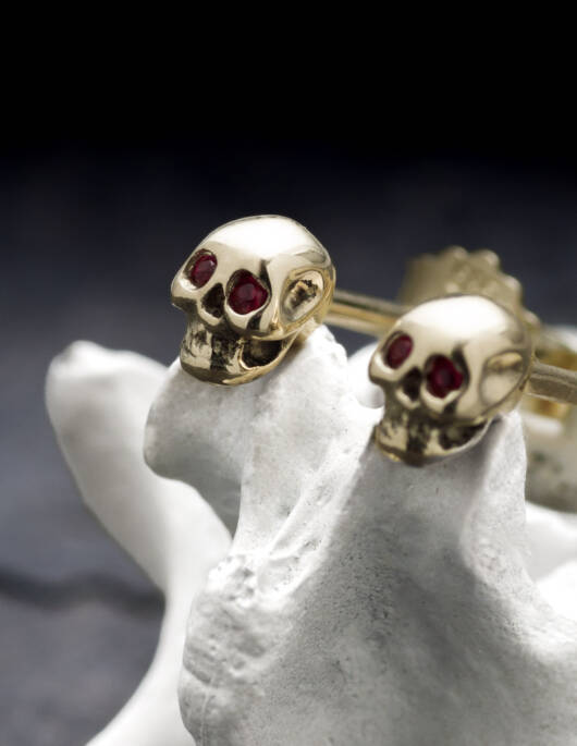 Detail of small skull earstuds made of yellow gold with ruby eyes.